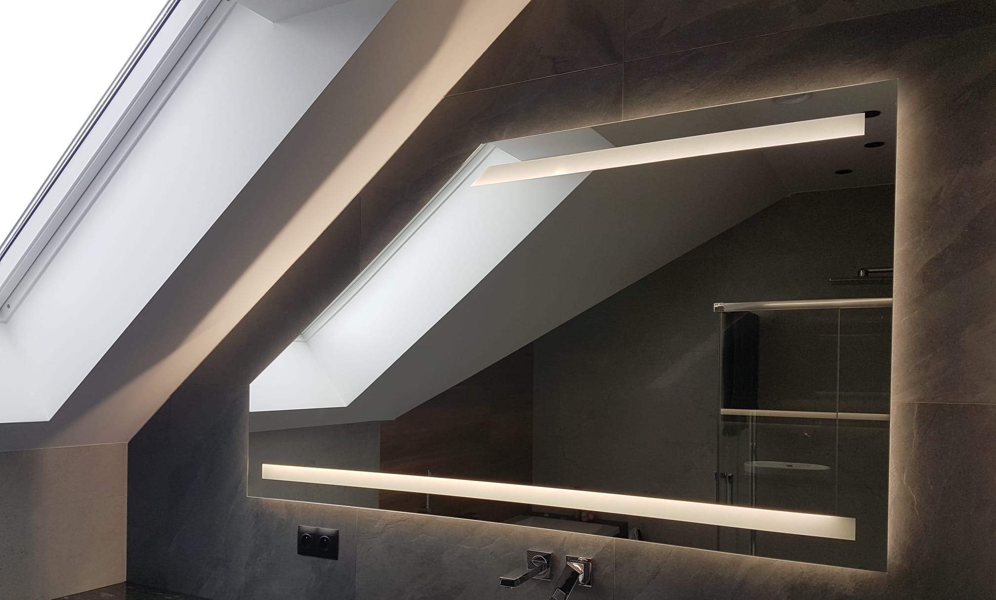 Oświetlone lustro w łazience na poddaszu, z widocznym oknem dachowym i szarą glazurą. Nowoczesny design, odbicie kabiny prysznicowej.