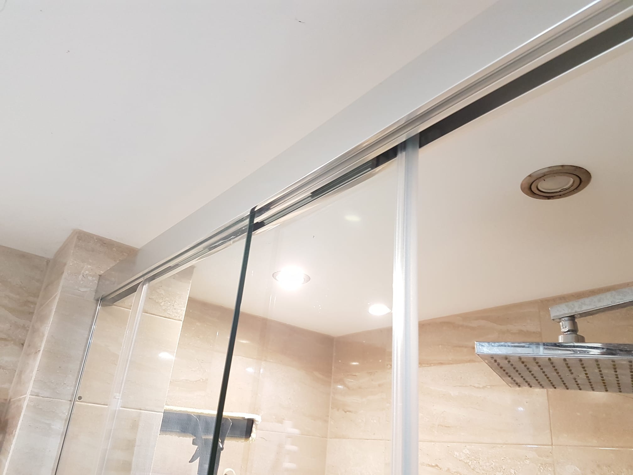 Szklana kabina prysznicowa z przesuwnymi drzwiami, zamontowana w łazience wykończonej beżowymi płytkami. Widoczna srebrna szyna i deszczownica.