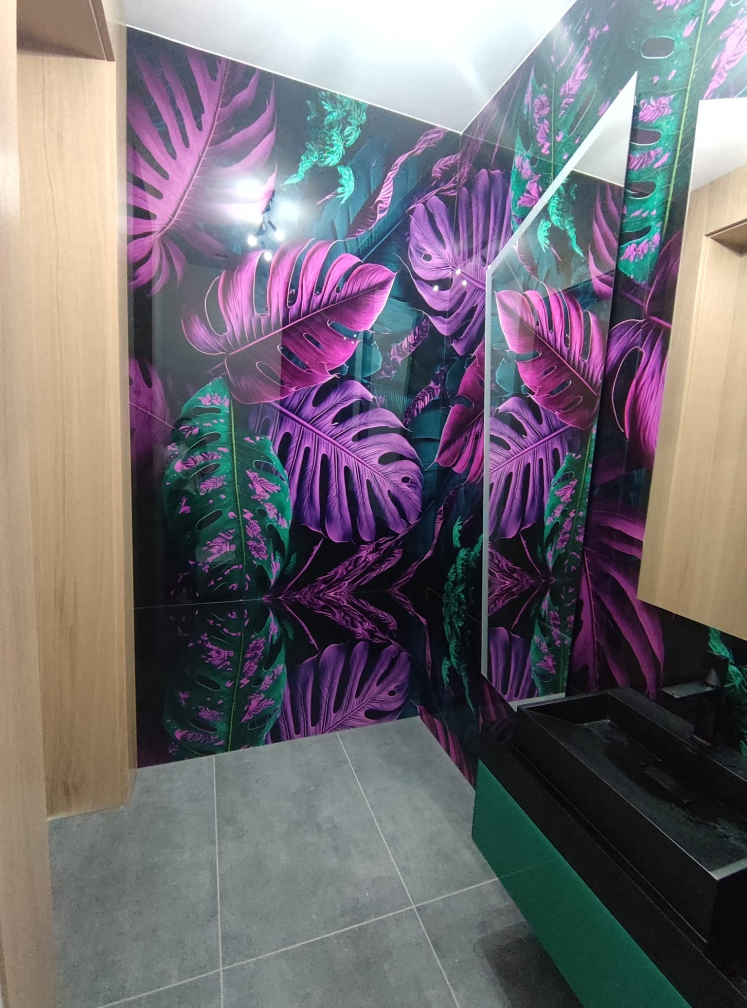 Szklana ściana z motywem tropikalnych liści w łazience, odbicie wzoru w połyskującej powierzchni. Nowoczesny design, szare płytki podłogowe, drewniane wykończenie.