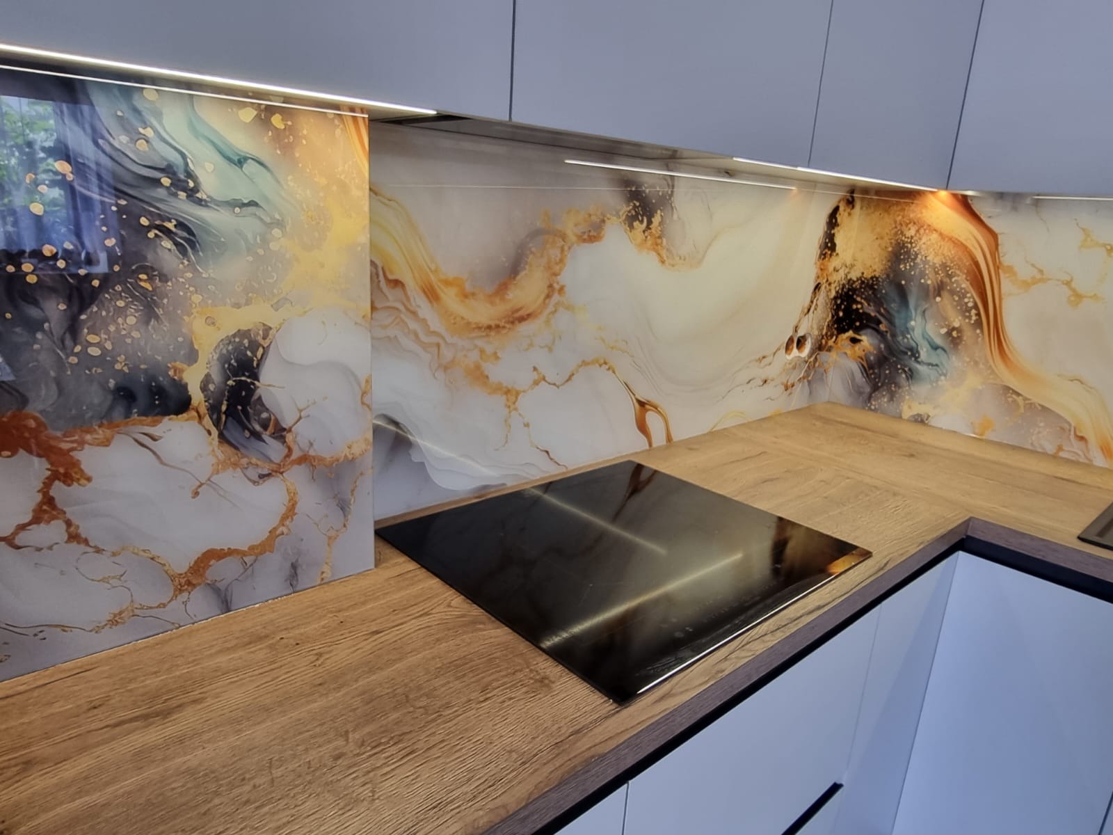 Szklany panel dekoracyjny z abstrakcyjnym wzorem, zamontowany nad blatem kuchennym z płytą indukcyjną, podświetlony listwą LED.