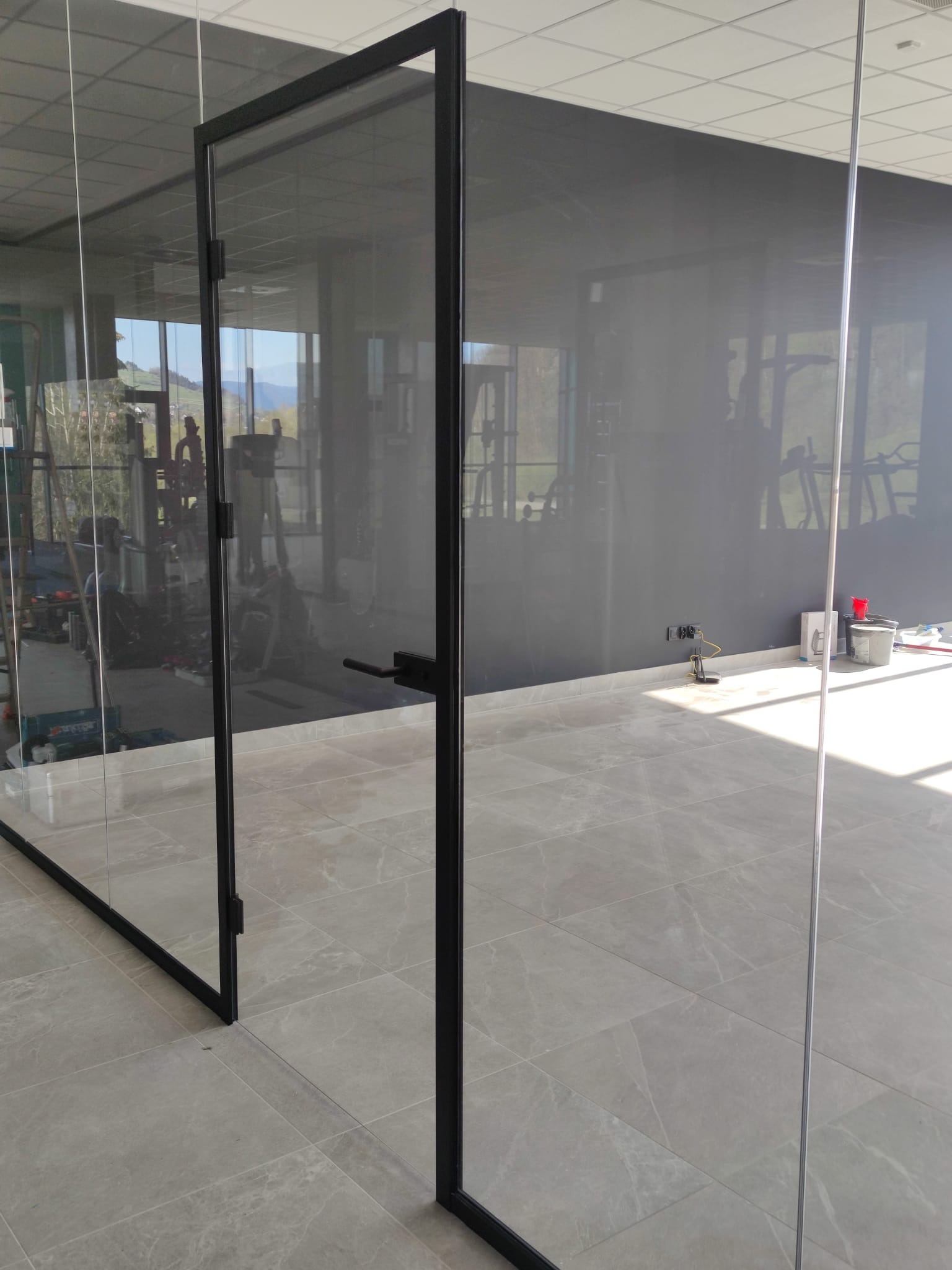 Szklane drzwi z czarną ramą, otwarte w nowoczesnym wnętrzu siłowni. Widoczne sprzęty do ćwiczeń i jasne płytki podłogowe.