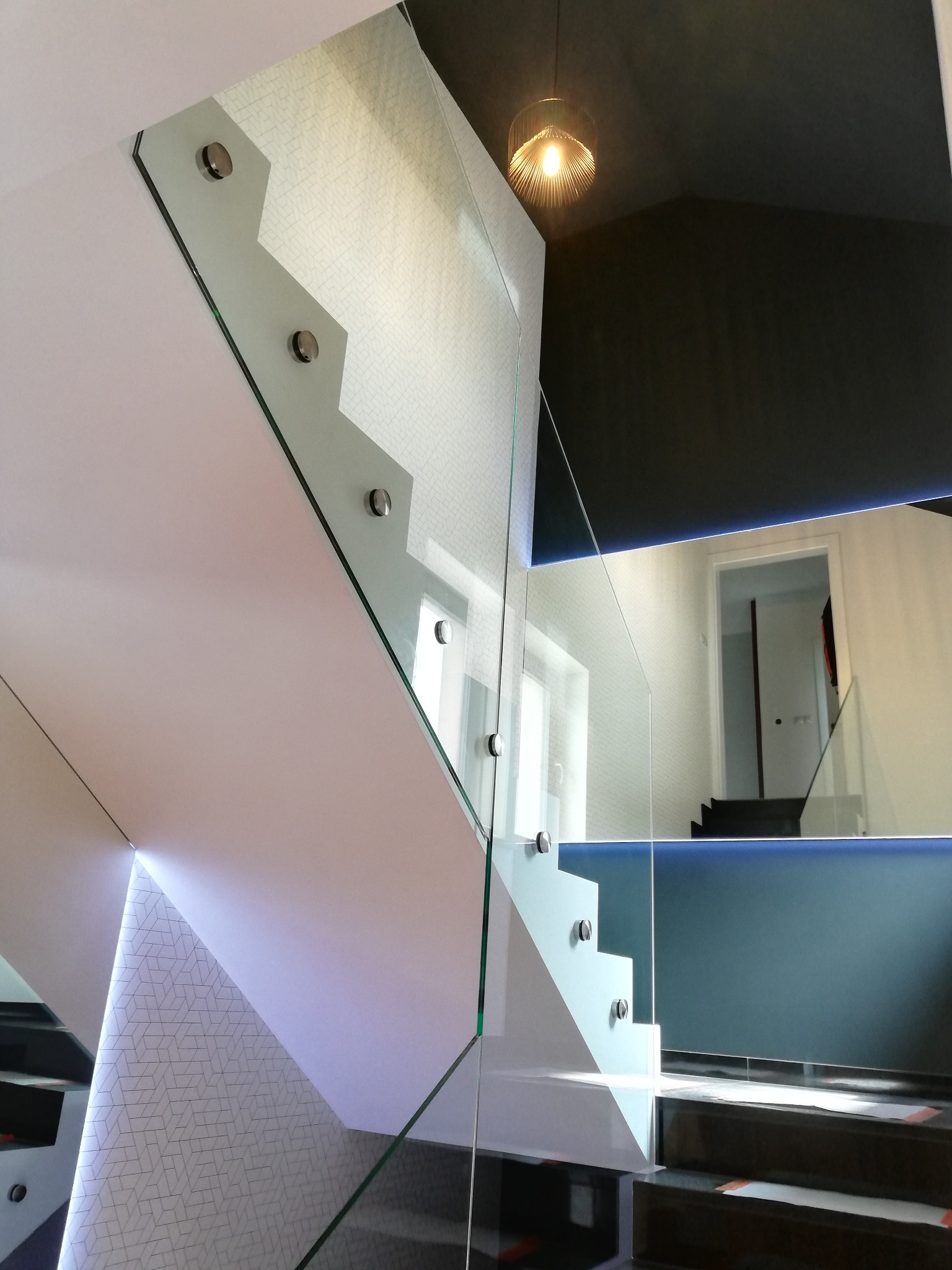 Nowoczesne schody z szklaną balustradą, mocowaną na metalowe uchwyty. Widoczne oświetlenie LED i lampa sufitowa, tworzące elegancki efekt wizualny.