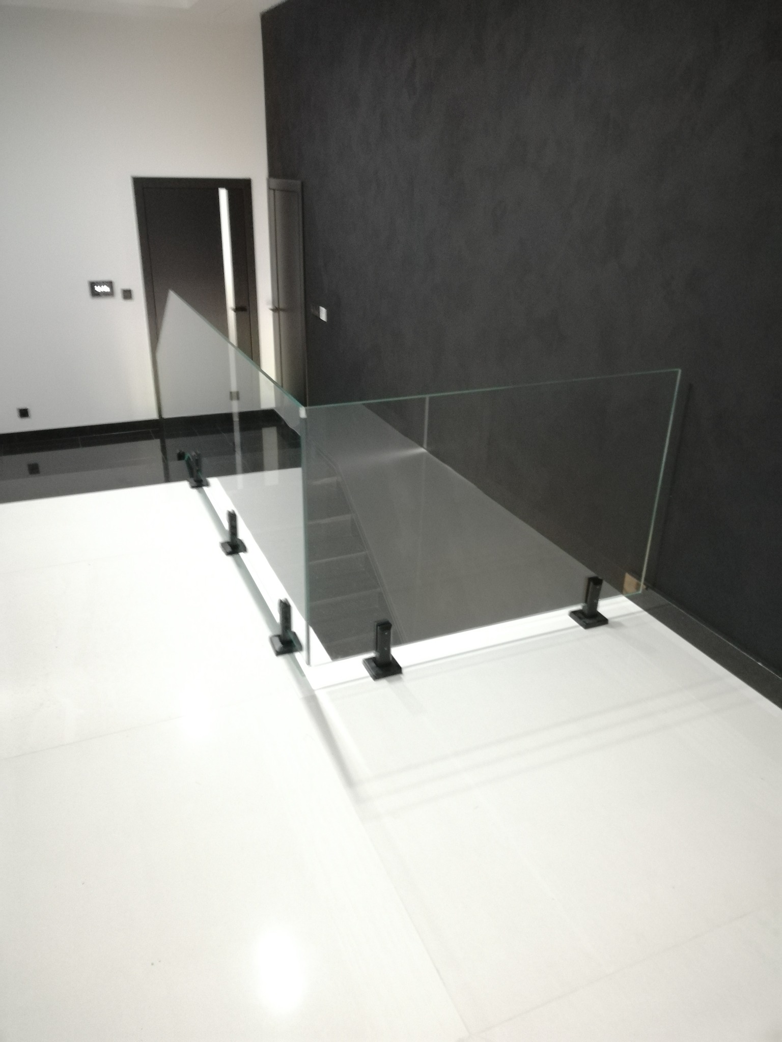 Nowoczesna balustrada ze szkła hartowanego, zamocowana na czarnych wspornikach, oddziela schody w minimalistycznym wnętrzu z białymi płytkami i ciemną ścianą.