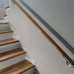 Drewniana poręcz ze stalowymi uchwytami zamontowana na białej ścianie wzdłuż drewnianych schodów.