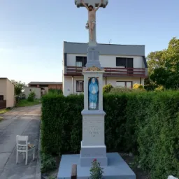 Odnowiony przydrożny krzyż z figurą Jezusa i Maryi, pomalowany na szaro-biało, na tle domu i zieleni.
