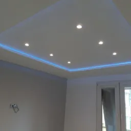 Podwieszany sufit z oświetleniem LED i punktowymi lampami w nowocześnie wykończonym wnętrzu, widok na szarą ścianę i okno.