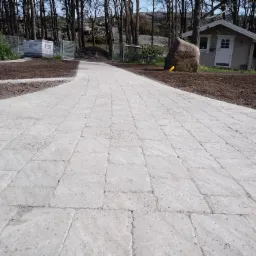 Świeżo ułożona, zakrzywiona ścieżka z jasnoszarej kostki brukowej prowadząca do domku narzędziowego, z widocznymi paletami kostki w tle i żółtą rurą drenażową przy trawniku.