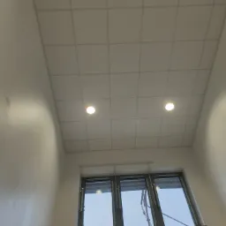 Widok z dołu na sufit podwieszany z widocznymi płytami, dwoma okrągłymi lampami i czujnikiem dymu, ściany pomalowane na biało.