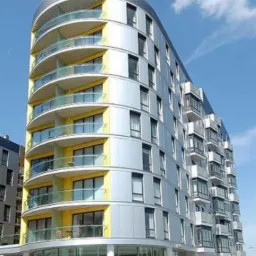 Nowoczesny, wielopiętrowy budynek mieszkalny z zaokrągloną elewacją, wykończony panelami w kolorze srebrnym i żółtym, z balkonami z przezroczystymi balustradami, sfotografowany w słoneczny dzień.