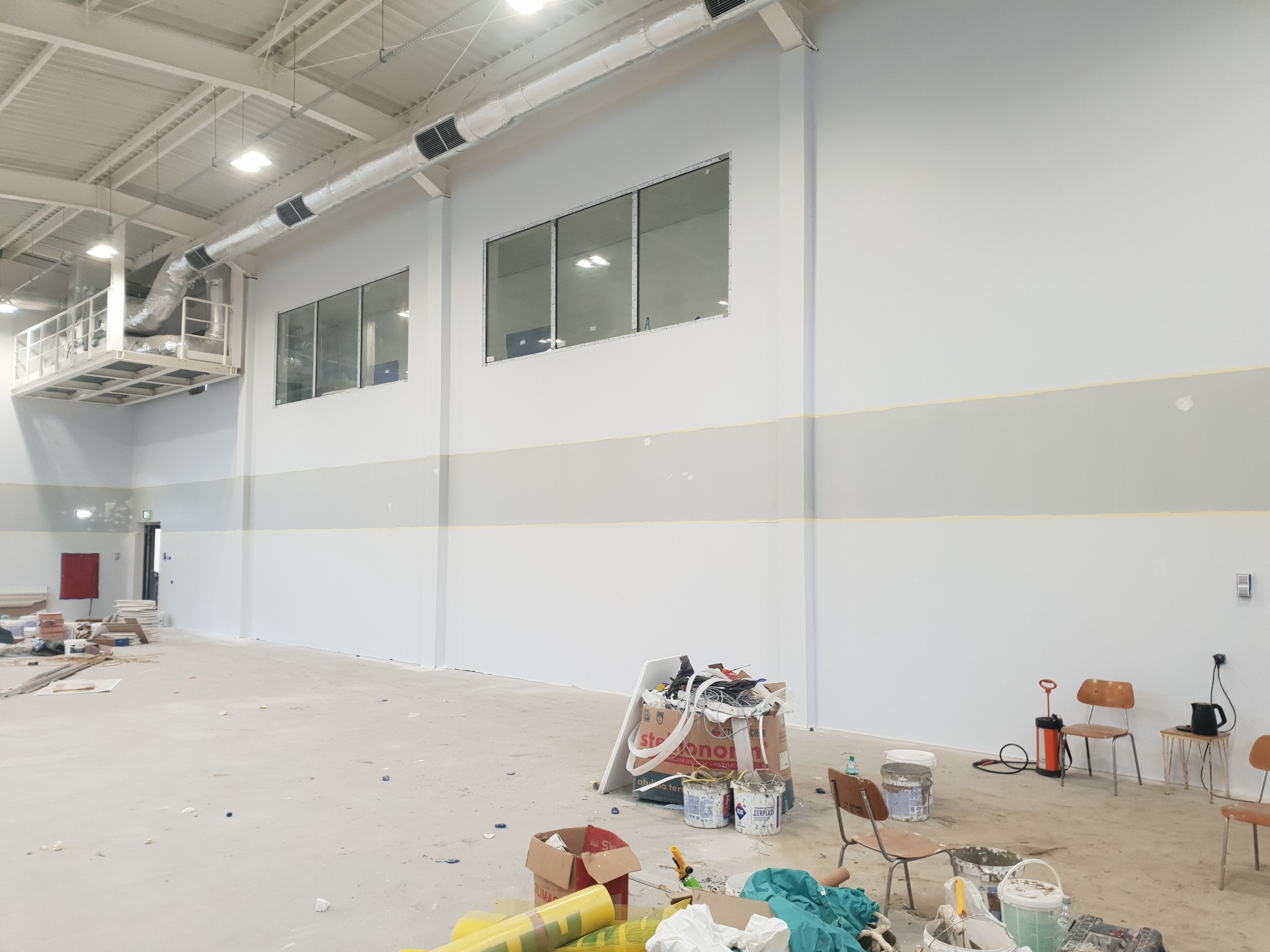 Wnętrze hali w trakcie remontu: pomalowane ściany na biało i szaro, widoczne okna, instalacja wentylacyjna pod sufitem, materiały budowlane i narzędzia malarskie porozrzucane na podłodze, krzesła...