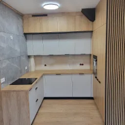 Nowoczesna kuchnia w trakcie montażu: szare szafki dolne i górne, drewnopodobne blaty i wykończenia, płyta indukcyjna w blacie, widoczne ślady po instalacji na ścianie nad blatem.