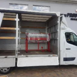 Biały samochód dostawczy z otwartą przestrzenią ładunkową, w której znajduje się czerwona maszyna do obróbki blach, zabezpieczona folią stretch. W tle widoczny budynek z logo firmy 'PROD-MASZ'.