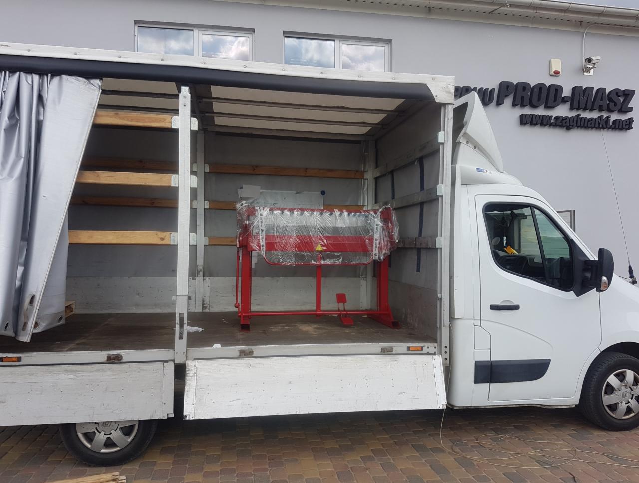 Biały samochód dostawczy z otwartą przestrzenią ładunkową, w której znajduje się czerwona maszyna do obróbki blach, zabezpieczona folią stretch. W tle widoczny budynek z logo firmy 'PROD-MASZ'.