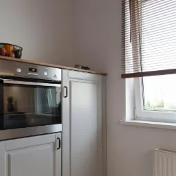 Kuchnia w jasnych barwach z zabudowaną srebrną kuchenką, drewnianym blatem, owocami w koszyku na szafce, drewnianymi żaluzjami w oknie i grzejnikiem pod oknem.