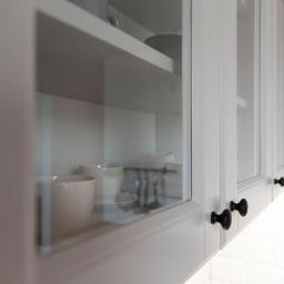 Janiecwnetrza - Szklane, białe szafki kuchenne z czarnymi gałkami, widoczne naczynia wewnątrz, oświetlenie LED pod szafkami, białe płytki na ścianie.