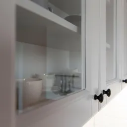 Szklane, białe szafki kuchenne z czarnymi gałkami, widoczne naczynia wewnątrz, oświetlenie LED pod szafkami, białe płytki na ścianie.