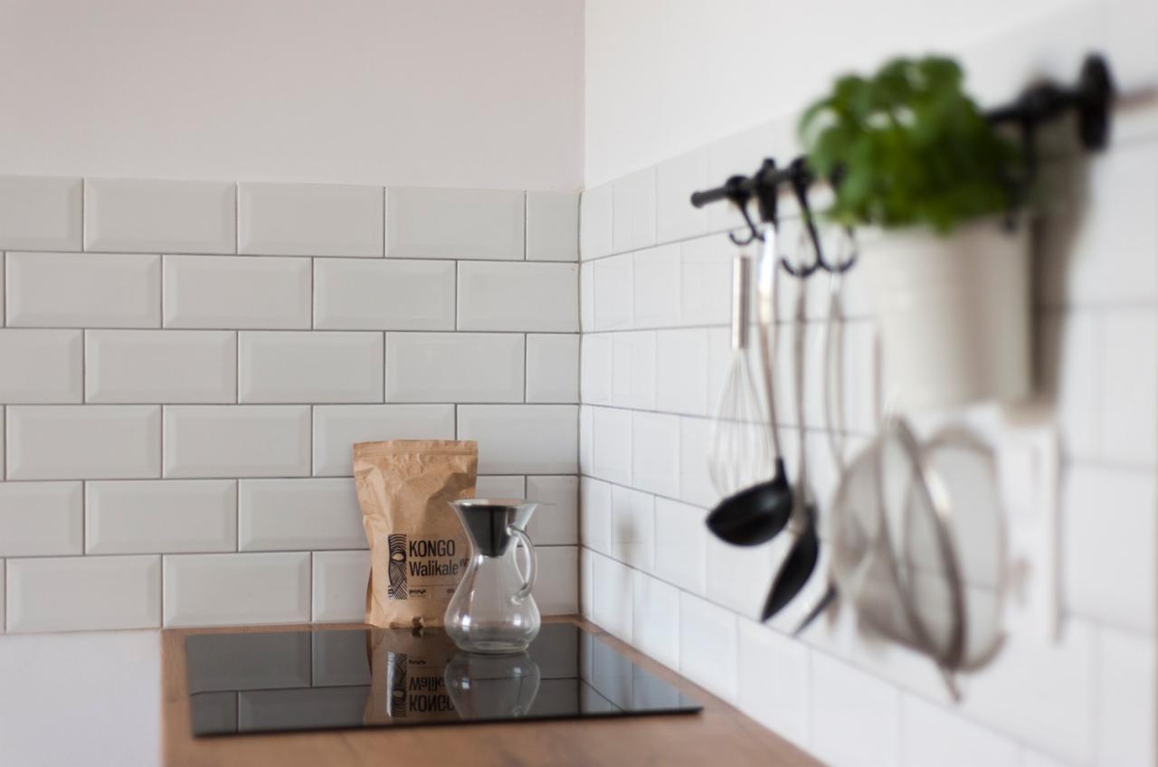 Minimalistyczna kuchnia z białymi, prostokątnymi płytkami na ścianie, brązowym blatem, szklanym dzbankiem do kawy i paczką kawy Kongo Walikale, z akcesoriami kuchennymi wiszącymi na relingu...