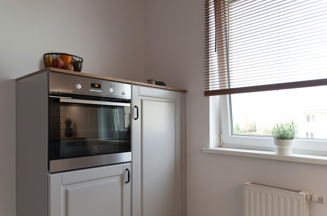 Kuchnia w jasnych barwach z zabudowaną srebrną kuchenką, drewnianym blatem, owocami w koszyku na szafce, drewnianymi żaluzjami w oknie i grzejnikiem pod oknem.