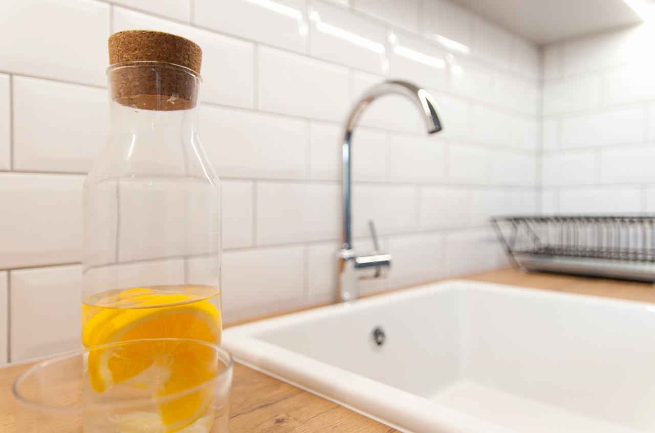 Biała, prostokątna umywalka w kuchni z baterią chromowaną, otoczona białymi, prostokątnymi płytkami. Na blacie stoi szklana butelka z korkiem, wypełniona wodą i plastrami pomarańczy, oraz szklanka.