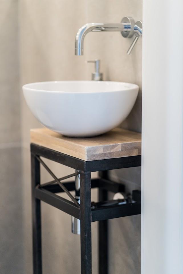 Biała umywalka nablatowa w kształcie misy na drewnianym blacie z metalową konstrukcją w stylu industrialnym, chromowana bateria ścienna, minimalistyczna łazienka.