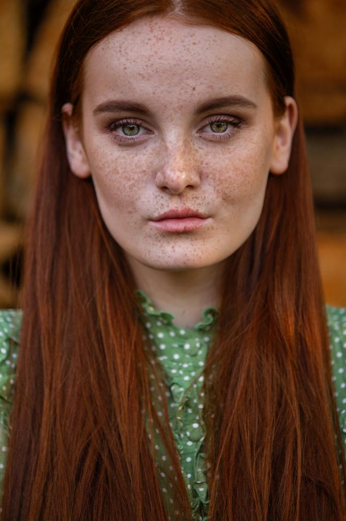 Portret młodej kobiety z rudymi włosami i zielonymi oczami, skupiającej wzrok w obiektyw, na tle drewnianej ściany.