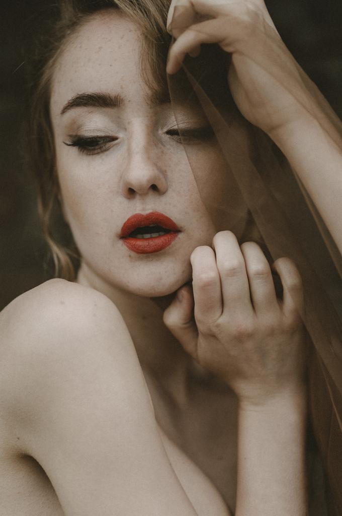 Portret kobiety z piegami, czerwonymi ustami i delikatnym makijażem oczu, częściowo zasłoniętej transparentnym materiałem.