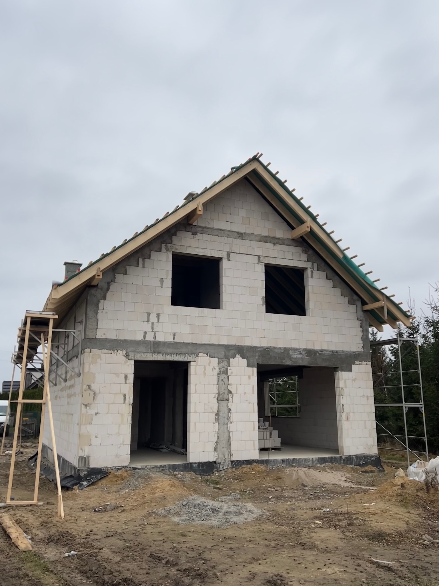 Budowa domu z białych bloczków: surowy stan, widoczne otwory okienne i drzwiowe, drewniana konstrukcja dachu. Na pierwszym planie ziemia i rusztowanie.