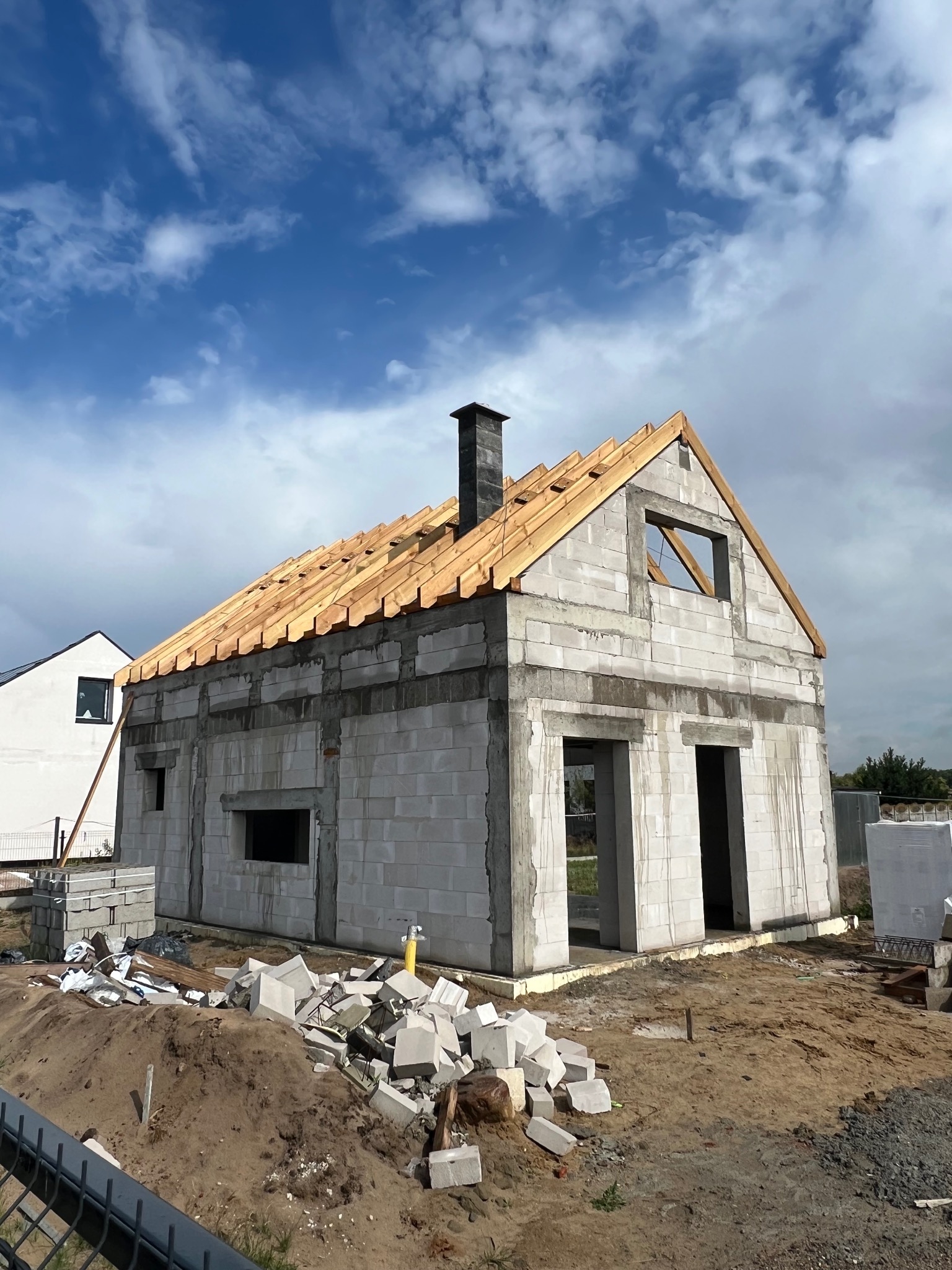 Surowy dom z pustaków z drewnianą konstrukcją dachu w trakcie budowy, widok z zewnątrz, w tle błękitne niebo z chmurami. Widoczne porozrzucane materiały budowlane.