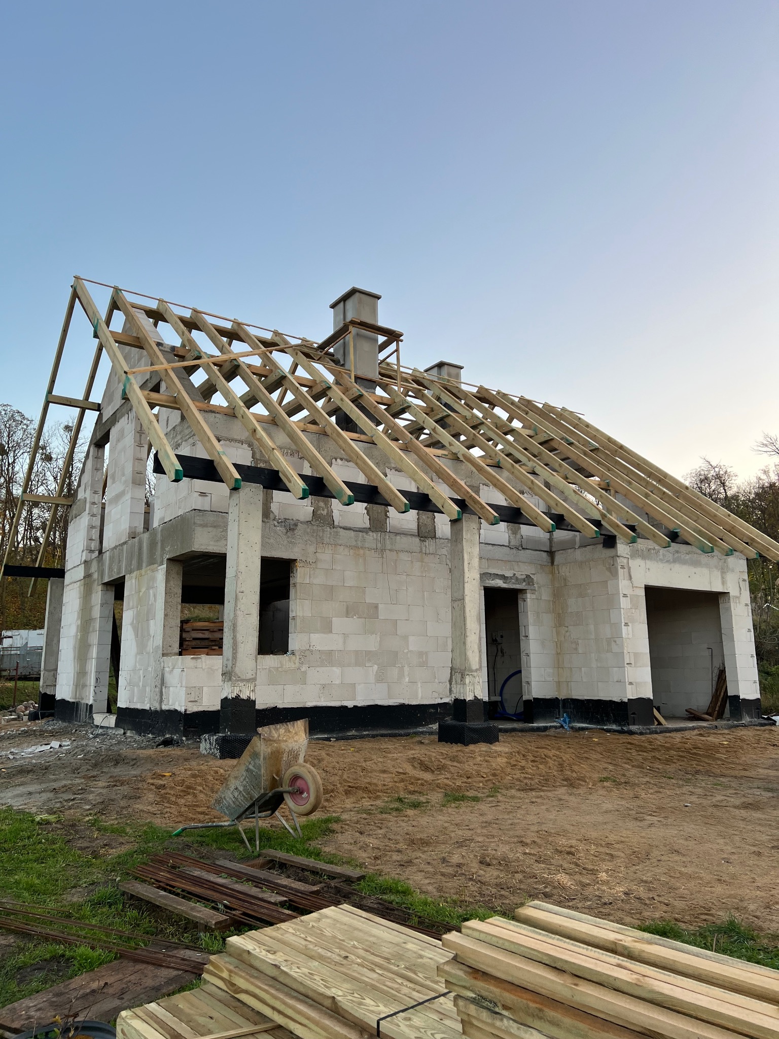 Budowa domu z bloczków: surowy stan z drewnianą konstrukcją dachu. Widoczna taczka i drewno na ziemi. Ujęcie z dołu, niebo w tle. Widuchowa 2025.