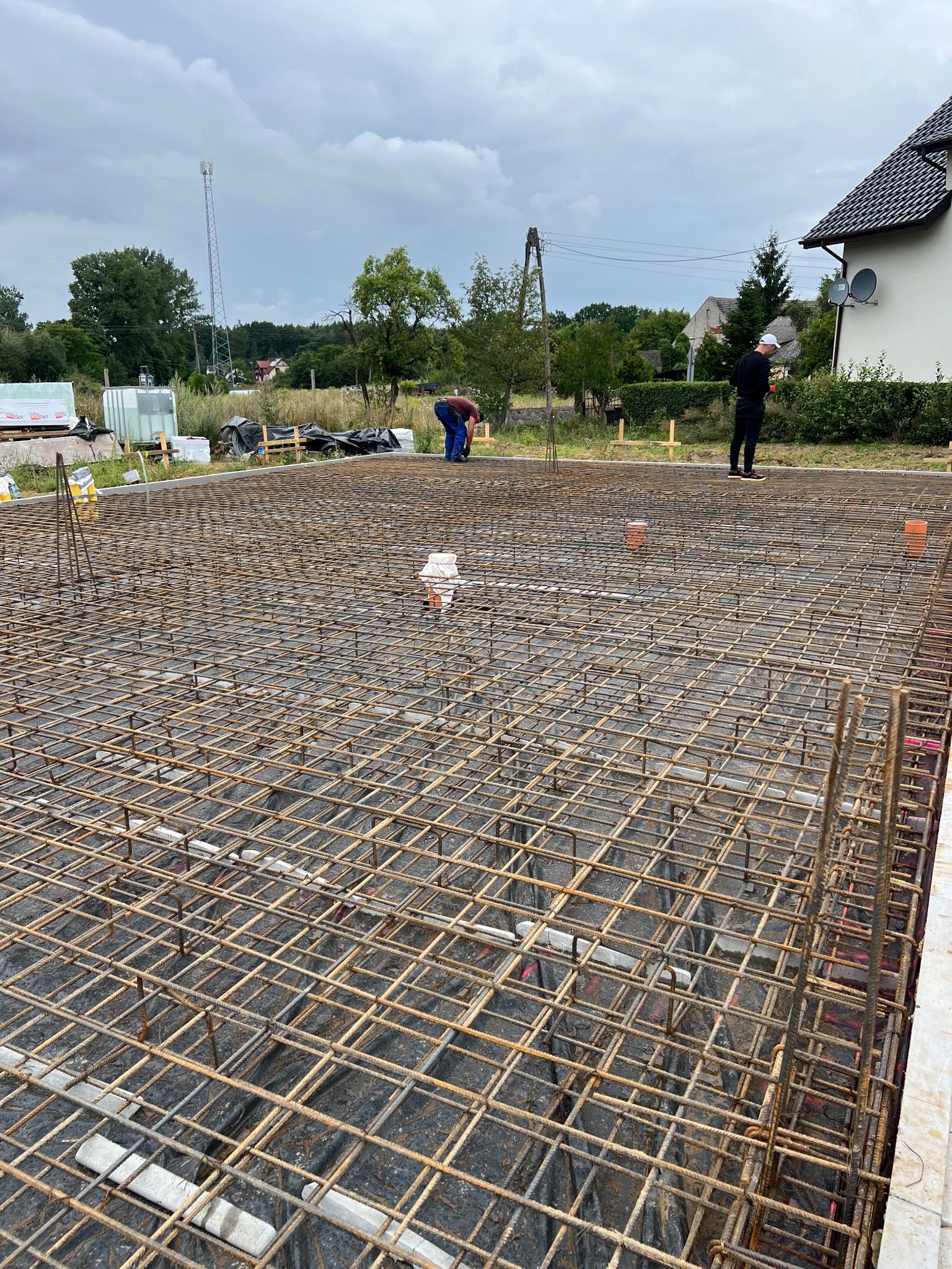 Zbrojenie fundamentu domu jednorodzinnego z widocznymi pracownikami w tle, wykonane z siatki stalowej na czarnej folii, Chwarstnica 2025.