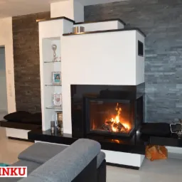 Nowoczesny salon z paleniskiem w obudowie z białych i czarnych płyt, płomienie tańczą za szybą, obok półki z dekoracjami i kamienna ściana.