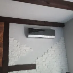 Szara klimatyzacja ścienna zamontowana w pomieszczeniu z dekoracyjną ścianą z białej cegły i drewnianymi belkami stropowymi.