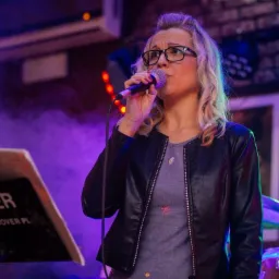 Wokalistka w okularach śpiewa do mikrofonu na scenie z perkusją w tle, oświetlona fioletowym światłem i z nutami na pulpicie z napisem 'COVER KAPELA-COVER.PL'.