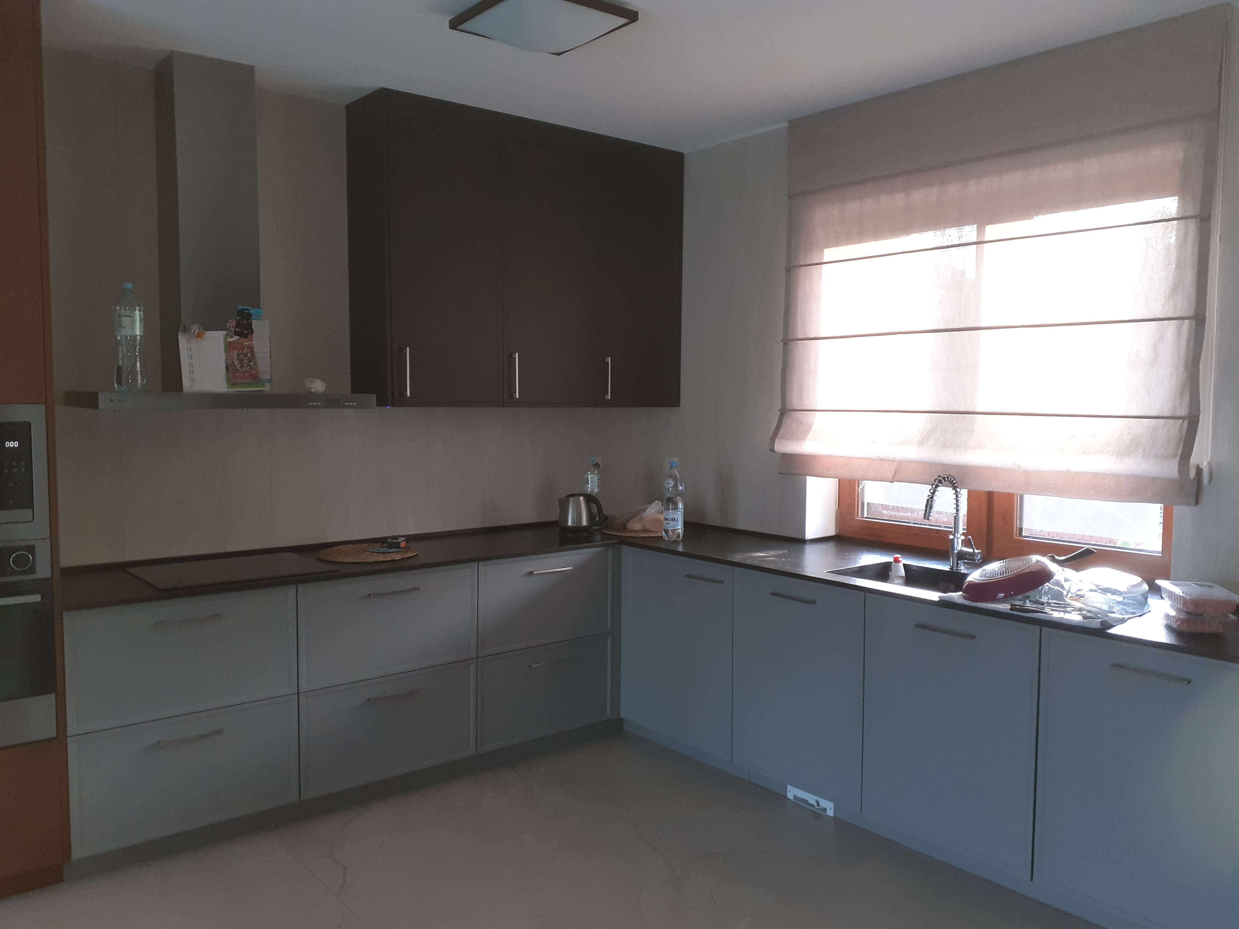 Nowoczesna kuchnia z szarymi szafkami, ciemnym blatem i brązowymi szafkami wiszącymi. W oknie widoczne rolety rzymskie. Na blacie zlewozmywak z naczyniami i butelki z wodą.