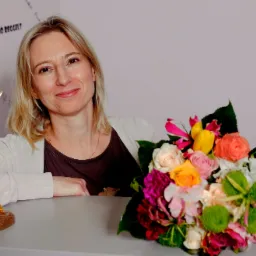 Portret uśmiechniętej kobiety z blond włosami, obok niej stoi statuetka i bukiet kolorowych kwiatów; na ścianie widoczne napisy w różnych językach.