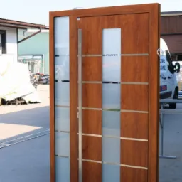 Brązowe drzwi wejściowe z przeszkleniami z matowego szkła, stalowy uchwyt, widoczny fragment budynku i samochodu dostawczego w tle.