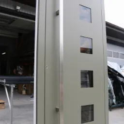 Nowoczesna brama garażowa segmentowa w kolorze szarozielonym z czterema kwadratowymi przeszkleniami i długim, pionowym uchwytem ze stali nierdzewnej, widoczna z boku na tle hali produkcyjnej.