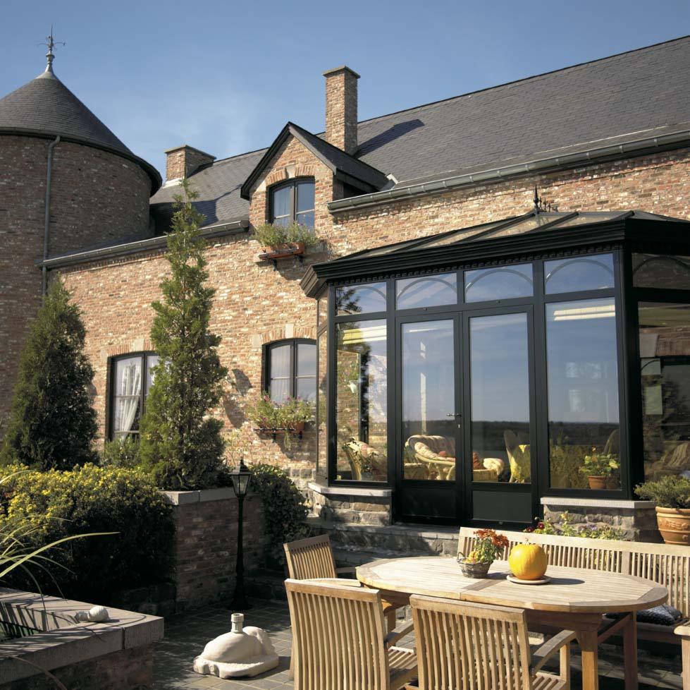 Szklana weranda z czarnymi ramami, przylegająca do ceglanego domu z wieżą, widok z tarasu z drewnianymi meblami ogrodowymi.