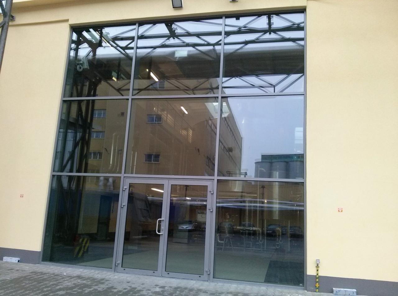 Duże przeszklenie z szarymi ramami i dwuskrzydłowymi drzwiami wejściowymi, odbijające budynek przemysłowy z metalowymi zbiornikami w tle.