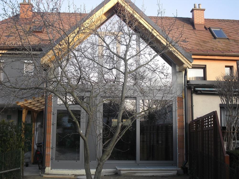 Rozbudowa domu z dużymi, przeszklonymi drzwiami przesuwnymi i oknami, widok z zewnątrz przez drzewo bez liści.