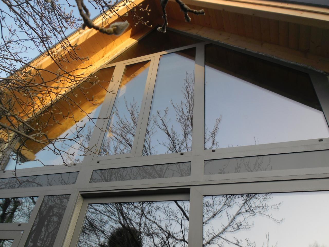 Duże, aluminiowe okno o nietypowym, trójkątnym kształcie, w szarej ramie, z widocznym drewnianym dachem i odbiciem drzew bez liści w szybach.