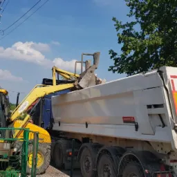 Żółta koparko-ładowarka B50 ładuje gruz do białej naczepy ciężarówki Langendorf na tle zieleni i błękitnego nieba z chmurami.