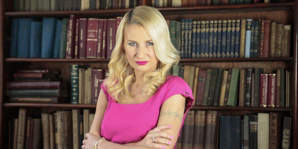 Portret kobiety w różowej sukience na tle biblioteczki z książkami, z tatuażem na ramieniu i pierścionkiem na palcu.