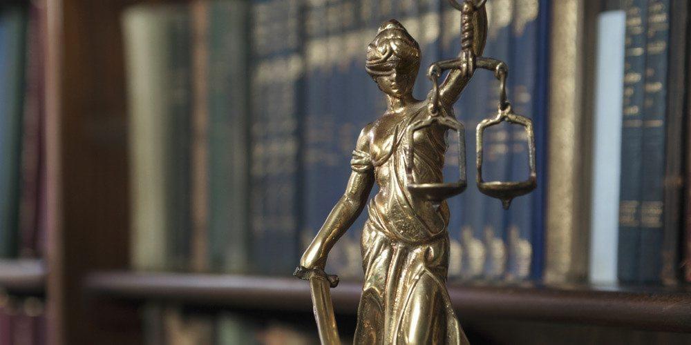 Złota statuetka Temidy, bogini sprawiedliwości z wagą i mieczem, na tle półki z książkami.