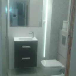 Wykończona łazienka z umywalką na szafce, toaletą podwieszaną i bidetem, oświetlenie LED pionowe oraz nad lustrem, szare płytki podłogowe i częściowo ścienne.