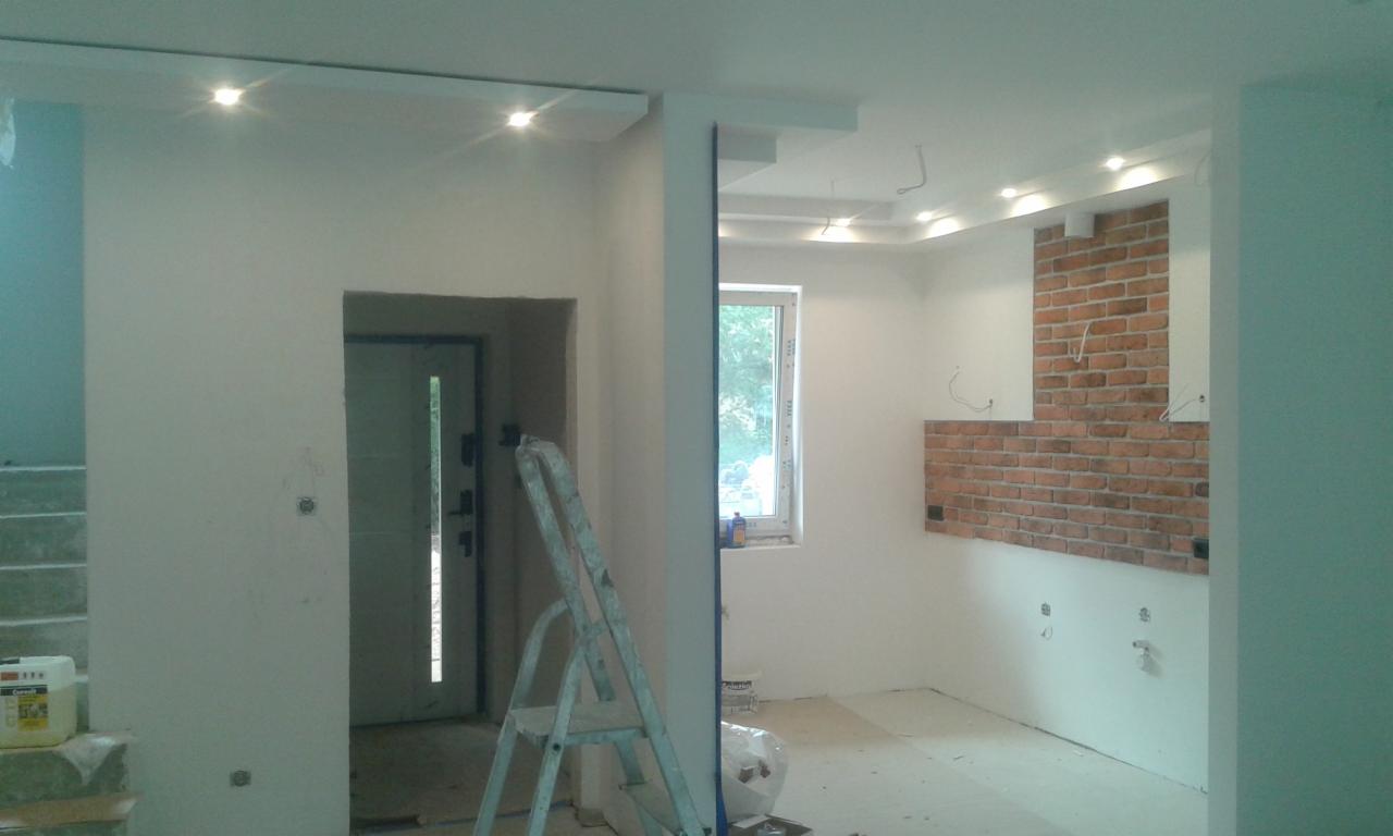 Wnętrze w trakcie prac wykończeniowych: białe ściany, fragment dekoracyjnej ściany z cegły, drabina oparta o ścianę przy drzwiach wejściowych, widoczne punkty oświetleniowe na suficie.