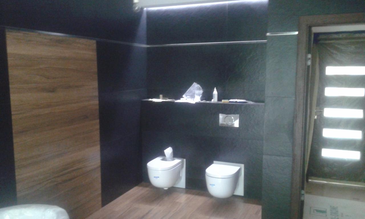 Nowoczesna łazienka z ciemnymi płytkami imitującymi kamień, drewnopodobną podłogą i wiszącymi toaletami, oświetlenie LED podsufitowe.