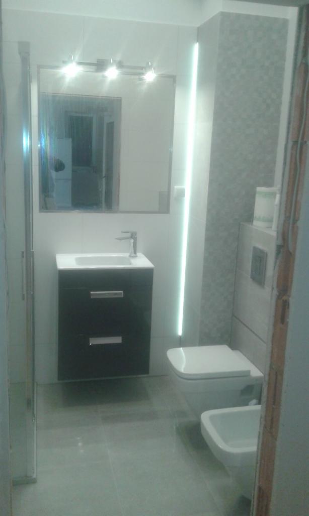 Wykończona łazienka z umywalką na szafce, toaletą podwieszaną i bidetem, oświetlenie LED pionowe oraz nad lustrem, szare płytki podłogowe i częściowo ścienne.