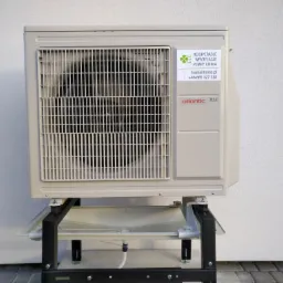 Kompaktowa zewnętrzna jednostka klimatyzacyjna Atlantic R32 na czarnej metalowej podstawie z tacką ociekową, zamontowana przy jasnej, teksturowanej ścianie.