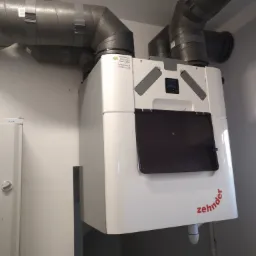 Biała centrala rekuperacyjna Zehnder z szarymi przewodami wentylacyjnymi w minimalistycznym pomieszczeniu, widoczny fragment okna i szafki.